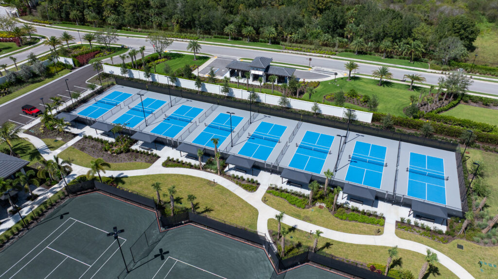 In-use lifestyle scene at the Caymas Naples sports center showing families playing pickleball on pristine courts, resort-style pool with waterslide in background, happy residents enjoying amenities, photorealistic 8K, vibrant and inviting